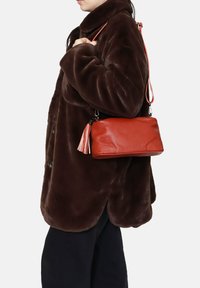 Borsa a mano rossa in pelle con una superficie testurizzata, dotata di una tracolla rosa removibile e un accento con nappa. Indossata con un cappotto marrone scuro.
