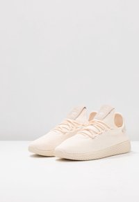 Par av ljusbeiga stickade sneakers med texturerade sulor och elastiska snören, placerade sida vid sida på vit yta.