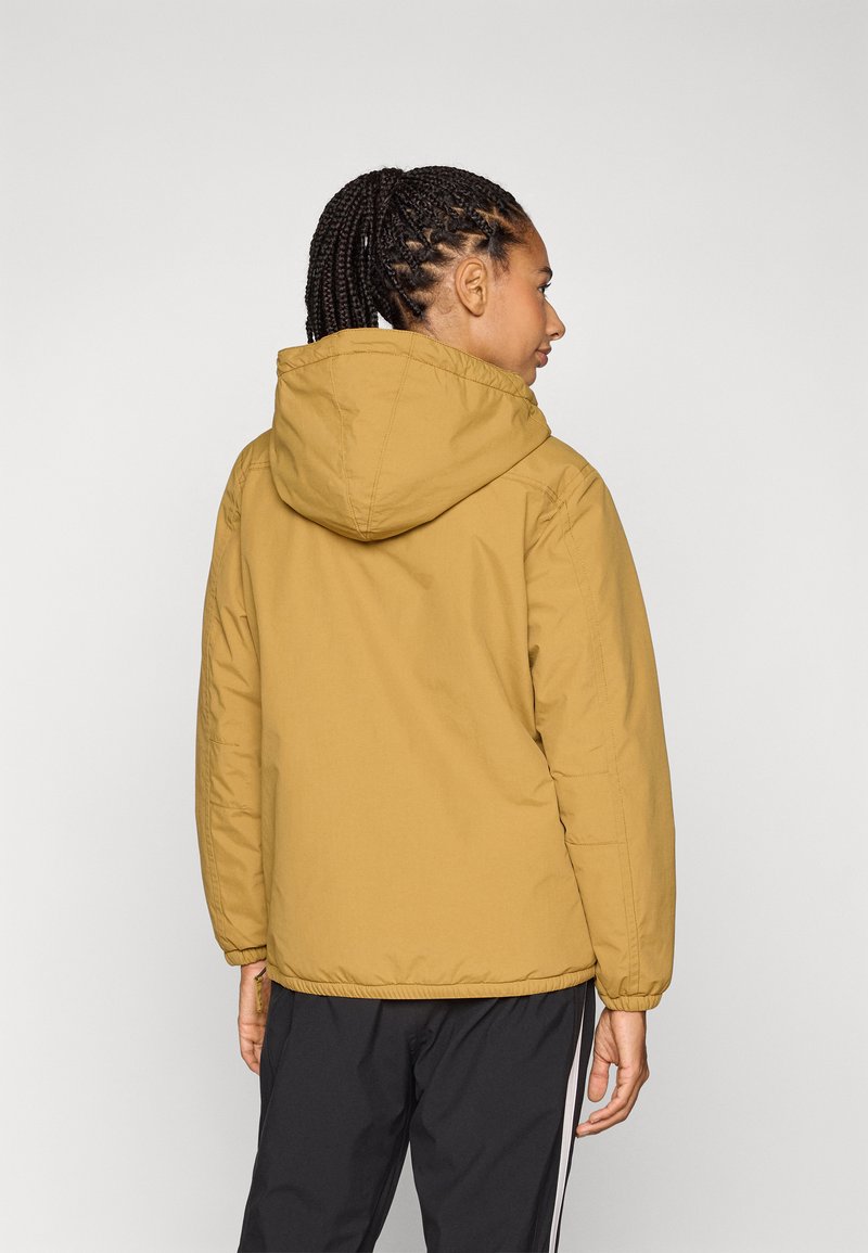 Gelbe gepolsterte Jacke mit Kapuze, die eine glatte Textur und elastische Bündchen hat. Das Model trägt schwarze Sporthosen.