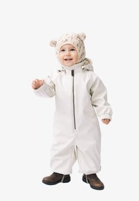 Combinaison de neige blanche imperméable avec fermeture éclair à l'avant, manches longues et poignets élastiques, accompagnée d'un bonnet beige en tricot avec des oreilles d'ours et de bottes marron.