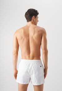 White swim shorts with an elastic waistband, rear pocket, and smooth fabric design, viewed from the back. Skin tone visible.