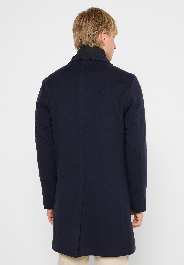 Manteau en laine mélangée bleu marine au coupe droite, à manches longues et à col tailleur. Design minimaliste, texture lisse et détail de fente au dos.