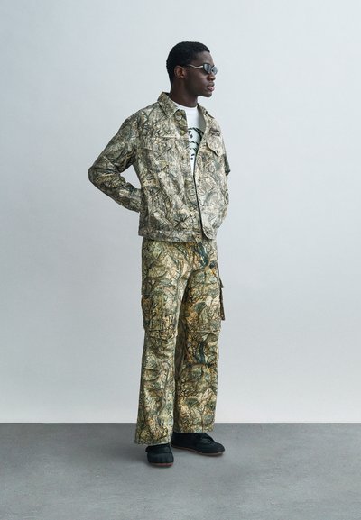 Τζάκετ καμουφλάζ και παντελόνι cargo σε τόνους πράσινου και καφέ με υφή σχεδίου. Το μοντέλο φοράει μαύρα παπούτσια και γυαλιά ηλίου.
