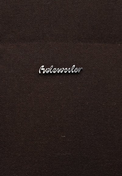 Metāla logotips "Holzweiler" rakstīts kursīvā, sudraba krāsā uz tumši brūnas teksturētas auduma fona. Logotips novietots centrā un izcelts.