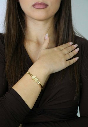 Pulsera de oro con motivos de mariposas y eslabones entrelazados, usada en la muñeca, con un acabado suave y pulido. El modelo tiene una postura neutral del torso superior.