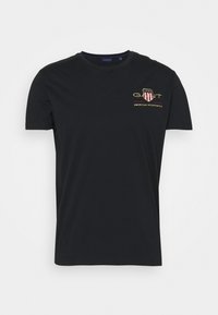 T-shirt noir à manches courtes avec petit logo poitrine représentant un bouclier, texte "GANT," et "American Sportswear" sur le côté gauche.