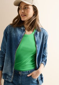 Giubbotto bomber in denim blu chiaro con zip frontale, colletto a costine e tasche laterali, abbinato a una maglietta senza maniche verde a coste.