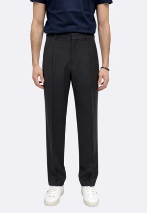 Uomo con pantaloni neri a pieghe, maglietta blu navy, sneakers bianche e braccialetto nero, in piedi con le braccia rilassate.