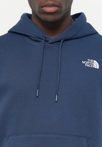 Marineblauwe hoodie met een voorzak in kangoeroestijl, koordjes en een wit logo op de linkerborst. Gemaakt van zachte stof.