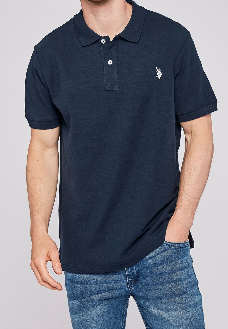 Homme portant un polo bleu marine avec deux boutons blancs et un petit logo brodé blanc sur la poitrine, associé à un jean bleu.