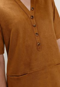 Hnědá košile s výstřihem do V, krátkými rukávy a pěti černými knoflíky. Obsahuje přední kapsu a měkkou, texturovanou tkaninu.