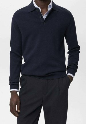 Mann trägt einen marineblauen Langarm-Polo-Pullover über einem weißen Hemd mit dunkler Hose, steht mit einer Hand in der Tasche.