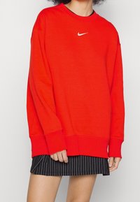 Rotes Sweatshirt mit gerippten Bündchen und Saum, versehen mit einem kleinen weißen Logo auf der Brust. Trägt man über einem schwarzen Minirock mit Nadelstreifen.