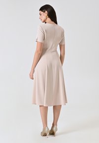 Hellrosa Kleid mit kurzen Ärmeln, enganliegendem Oberteil und ausgestelltem Rock. Mit einem Reißverschluss hinten und glattem, dehnbarem Material. Kombiniert mit goldenen High Heels.