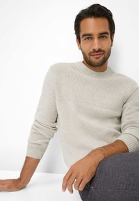Helles graues Sweatshirt mit einem strukturierten oberen Panel, rundem Ausschnitt und gerippten Bündchen. Das Modell hat schulterlanges dunkles Haar und sitzt.