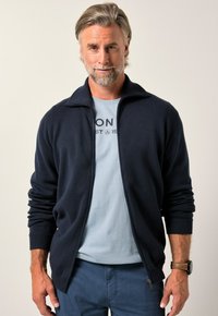 Giacca blu navy con zip, polsini e colletto a costine, realizzata in materiale morbido, indossata sopra una t-shirt azzurro chiaro con testo stampato.