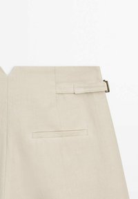 Jupe beige taille haute avec une sangle à boucle latérale et une poche arrière horizontale, présentée sur un fond blanc.