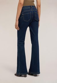 Osoba oblečená v tmavě modrých zvonových džínách a hnědých botách na podpatku stojící zády ke kameře na jednobarevném pozadí.