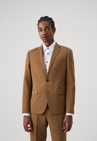 Browns uzvalks ar slim fit griezumu. Divas priekšējās kabatas, viens pogu aizdare. Kombinēts ar baltu krekls ar melnām pogām un aprocēm.