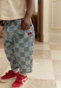 Pantalons à carreaux bleu clair avec un motif de cerise brodé et des baskets rouges avec des bandes auto-agrippantes sur un sol carrelé.