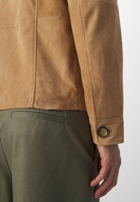 Gros plan d'une personne portant une veste en suède beige avec des poignets boutonnés et un pantalon vert olive, montrant le bas du dos et le bras droit.