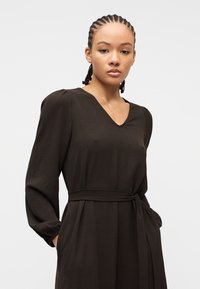 Νεαρή γυναίκα με πλεξούδες, φορώντας μακρυμάνικο μαύρο φόρεμα με V λαιμόκοψη και ζώνη στη μέση, στέκεται με τα χέρια στις τσέπες μπροστά σε απλό φόντο.