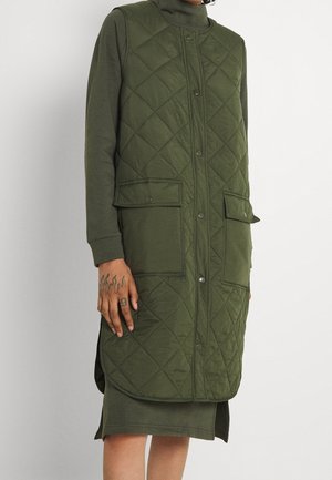 Hihaton tikattu oliivinvihreä liivi, jossa edessä läpälliset nappikiinnitykset ja kaksi taskua, puettu pitkähihaisen oliivinvihreän mekon päälle.