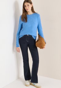 Hellblauer Pullover, dunkel ausgestellte Jeans, beigefarbene Wildledertasche und cremefarbene Sneakers mit blauen Akzenten. Glatte Texturen und kontrastierende Nahtdetails.