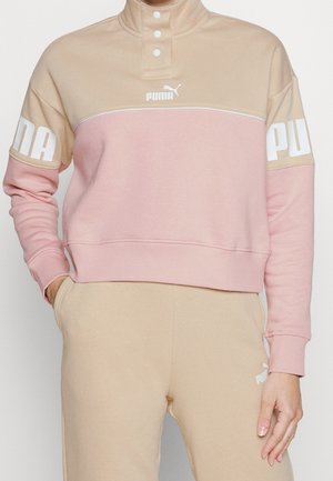 Sweat-shirt Puma bicolore beige et rose avec col à boutons-pression, logo sur la poitrine et les manches, assorti à un pantalon de survêtement beige Puma.