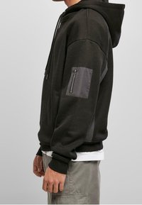 Vista lateral de una persona que lleva una sudadera con capucha negra y un bolsillo con cremallera en la manga, pantalones cargo grises y una camiseta blanca debajo.