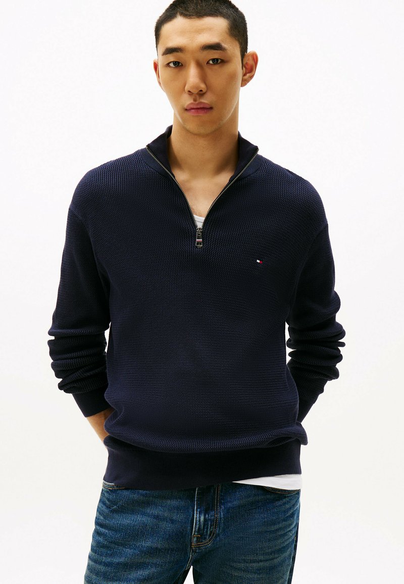 Pull-over en tricot bleu marine avec un col zippé, des manches longues et des poignets côtelés. Porté sur un T-shirt blanc, associé à un jean en denim bleu.