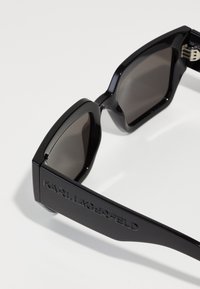 Lunettes de soleil noires avec une monture épaisse et angulaire, des verres sombres, et un logo "KARL LAGERFELD" embossé sur les branches. Texture lisse sur l'ensemble.