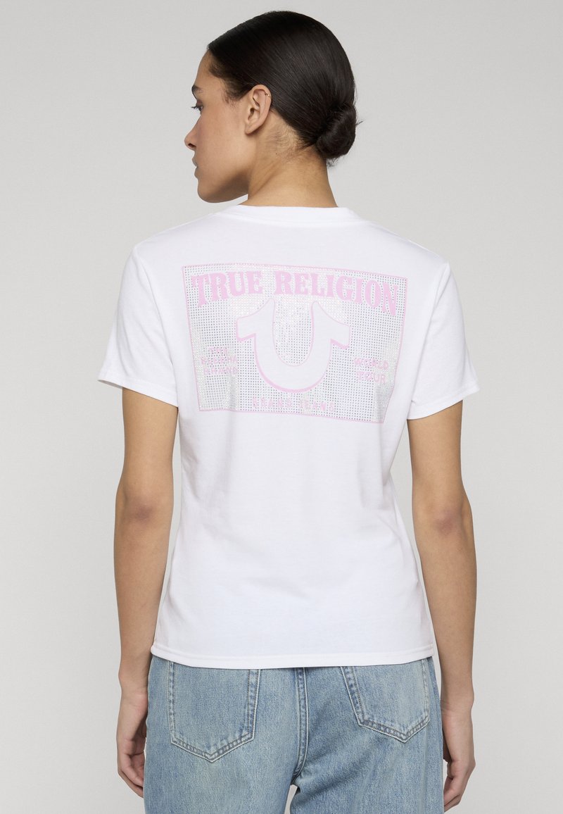 Femme aux cheveux foncés attachés en chignon, portant un t-shirt blanc True Religion avec un logo rose et un jean bleu clair, vue de dos sur un fond uni.