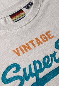 Lichtgrijze katoenen t-shirt met in oranje het woord "VINTAGE" en in gestructureerde teal letters "Superdry", met een ronde halsontwerp.