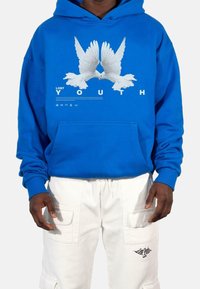 Modrá mikina s kapucí a bílou grafikou dvou holubic, pod níž je text "ZTRACENÁ MLADISTVOST". Obsahuje klokaní kapsu a žebrované manžety. Bílé cargo kalhoty.