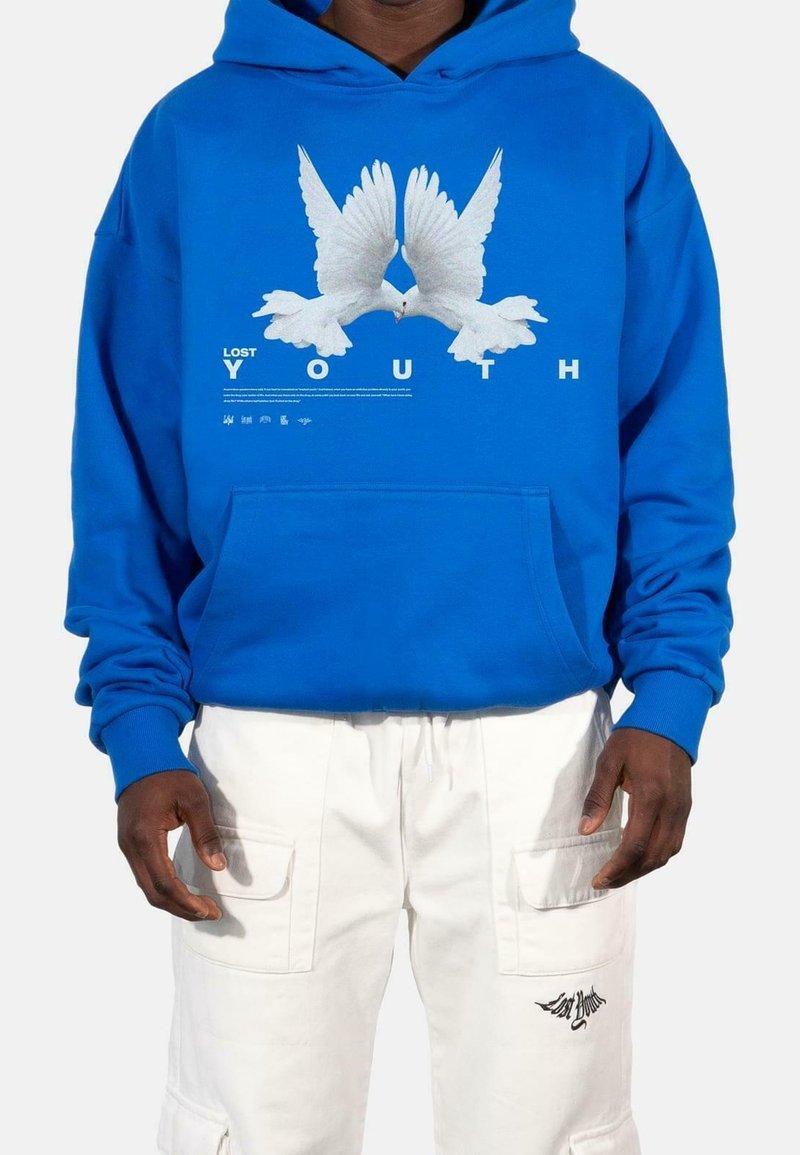 Modrá mikina s kapucí a bílou grafikou dvou holubic, pod níž je text "ZTRACENÁ MLADISTVOST". Obsahuje klokaní kapsu a žebrované manžety. Bílé cargo kalhoty.
