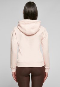 Lys rosa hettegenser med en koselig hette og lange ermer, med ribbestrikket kant og mansjetter, parret med brune leggings. Myk stofftekstur.