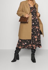 Brun ullblandad kappa över en svart blommig klänning med röda och rosa accenter. Svarta läderknähöga stövlar. Brun texturerad axelväska.