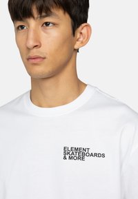 Hvid bomulds t-shirt med et sort trykt logo, der står "ELEMENT SKATEBOARDS & MORE" på venstre brystområde.