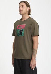 T-shirt à manches courtes en coton vert olive avec un design graphique coloré représentant des figures sur un fond rose et vert.