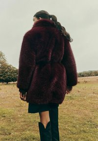 Burgundijas mākslīgā kažokāda pārklājs ar plašu apkakli un jostu viduklī, ar maigu tekstūru un pārspīlētu piegriezumu, valkāts pār tumšiem svārkiem un zābakiem.