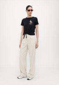 Svart bomull t-skjorte med knyttet hem, med et gullflamingo-grafikk. Kombinert med beige lin vid bukser og sneakers.