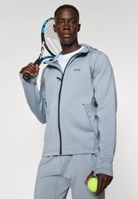 Haut de sweat à capuche zippé gris clair avec capuche, zipper noir et un branding subtil. Le modèle tient une raquette de tennis et une balle de tennis verte. Texture lisse.
