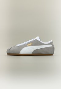 Γκρι σουέτ αθλητικό παπούτσι Puma με λευκή ρίγα, λευκά κορδόνια και καουτσούκ σόλα, εμφανιζόμενο σε προφίλ σε ουδέτερο φόντο.