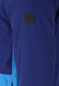 Blå och lila jacka med en mjuk textur, med en svart cirkulär logotyp på vänster ärm och kontrasterande ljusblå detaljer.