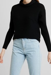 Černý žebrovaný pletený svetr s vysokým límcem a dlouhými rukávy, doplněný světle modrými džínami s vysokým pasem. Na svetru je patrná textura.