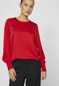 Blusa di raso rosso con maniche lunghe, caratterizzata da polsini texturizzati arricciati. Tessuto liscio e scollatura tonda, abbinata a pantaloni neri.