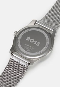 Silverfärgad klockbak i rostfritt stål med en jämn yta, meshband och graverad "BOSS"-logotyp. Vattentålig upp till 3 bar.