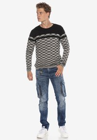 Schwarzer und grauer gemusterter Pullover mit einem Diamantdesign, kombiniert mit blauen Cargo-Jeans mit Reißverschlusstaschen und weißen Turnschuhen.