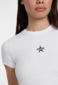 Valkoinen lyhythihainen t-paita, jossa on musta brodeerattu tähti ja "GUESS JEANS" -logo rinnassa. Sujuva puuvillakangas. Tiukka malli.